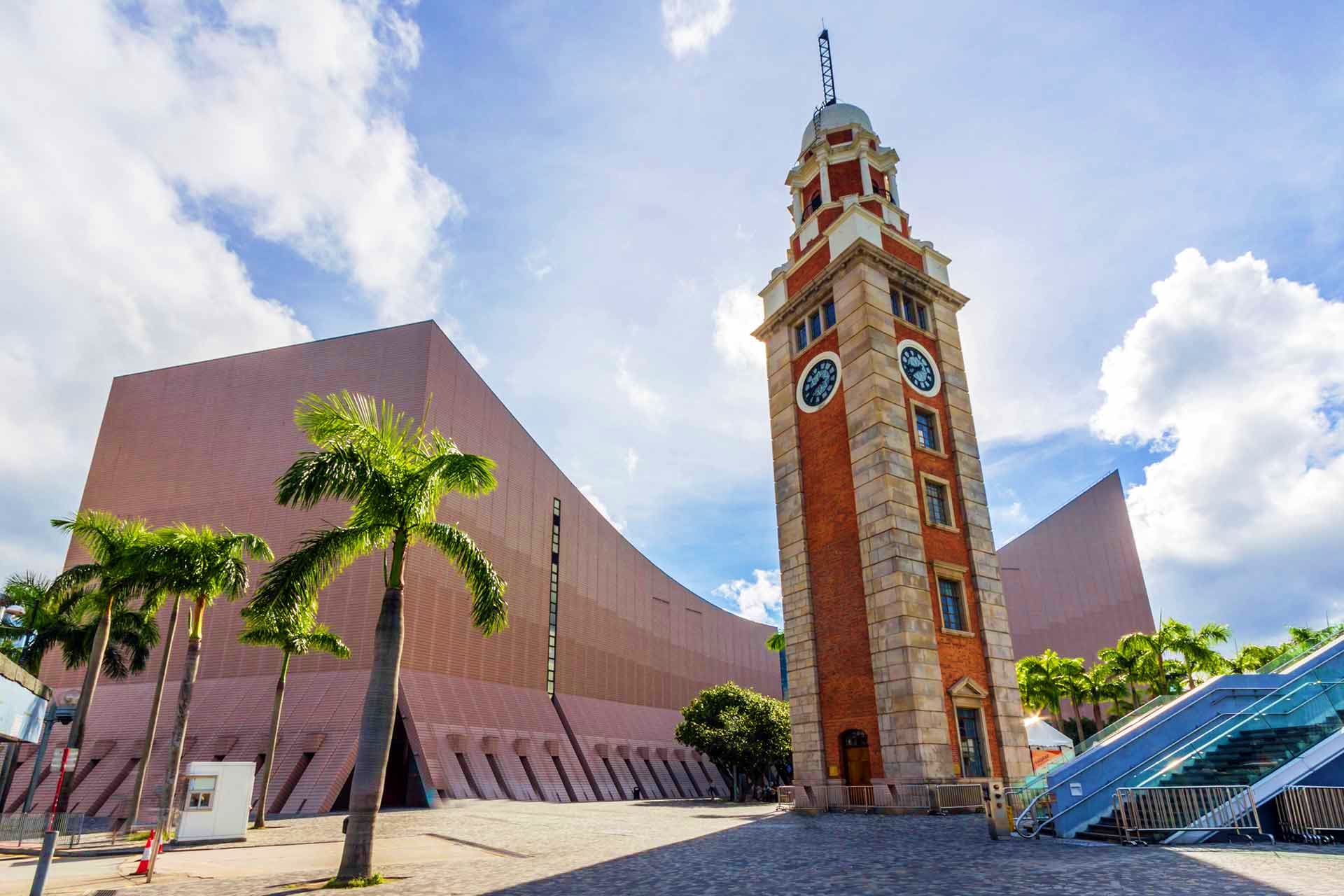 Historic red brick clock tower and modern museum building in Hong Kong waterfront plaza with palm trees